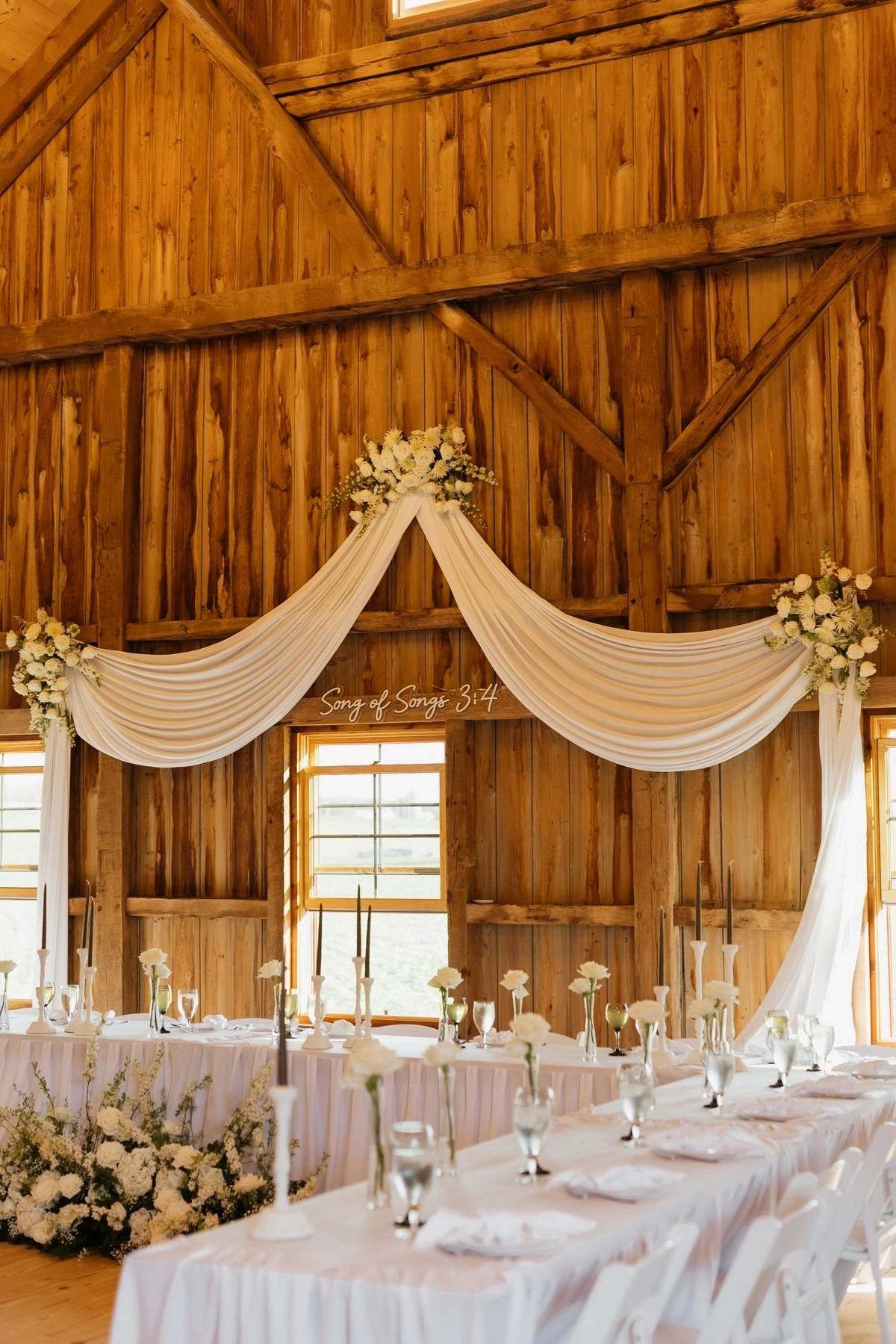 Barn interior head table with draped fabric, candles and white floral arrangements