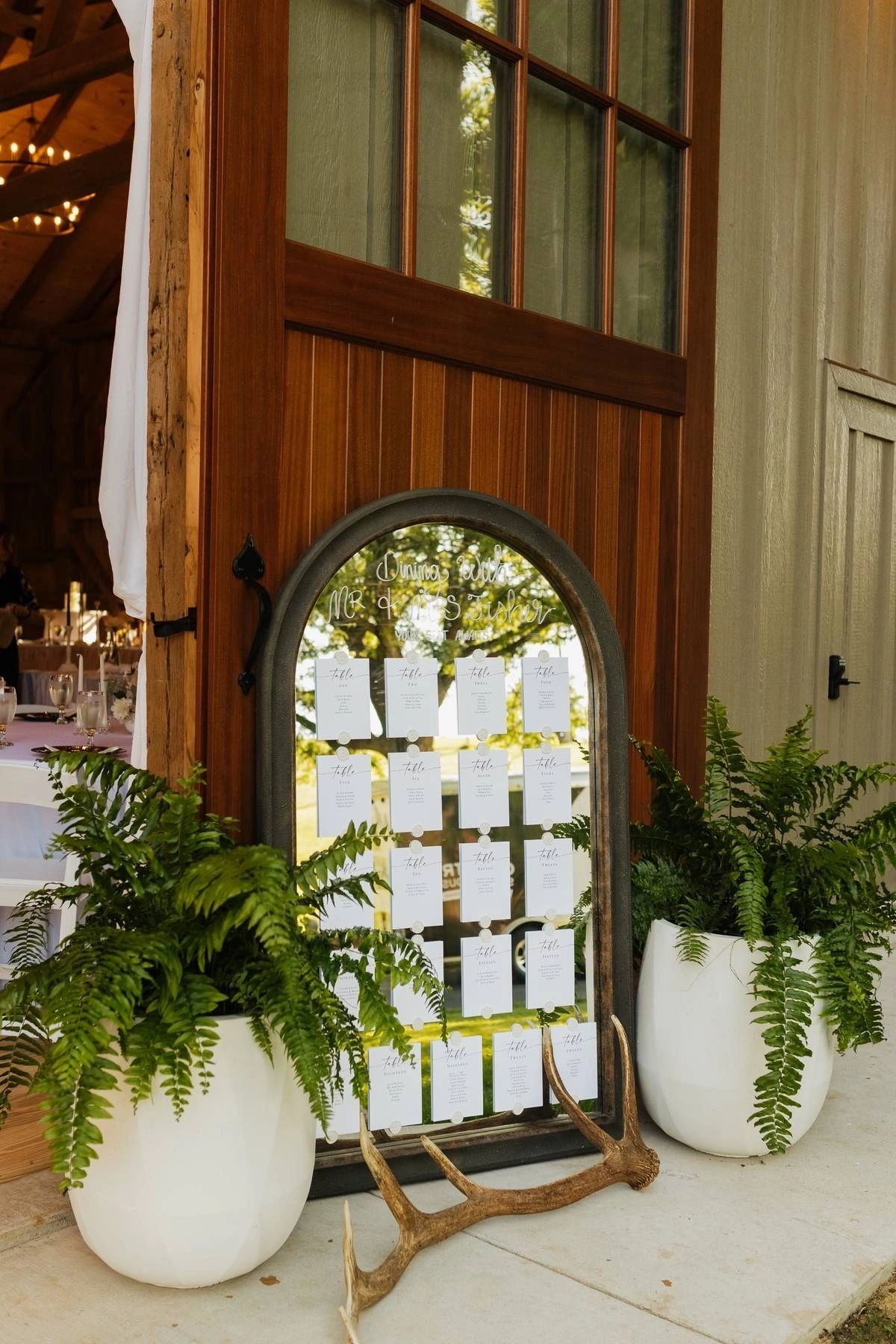 Entry vignette with arched mirror seating chart and potted ferns at the venue doors