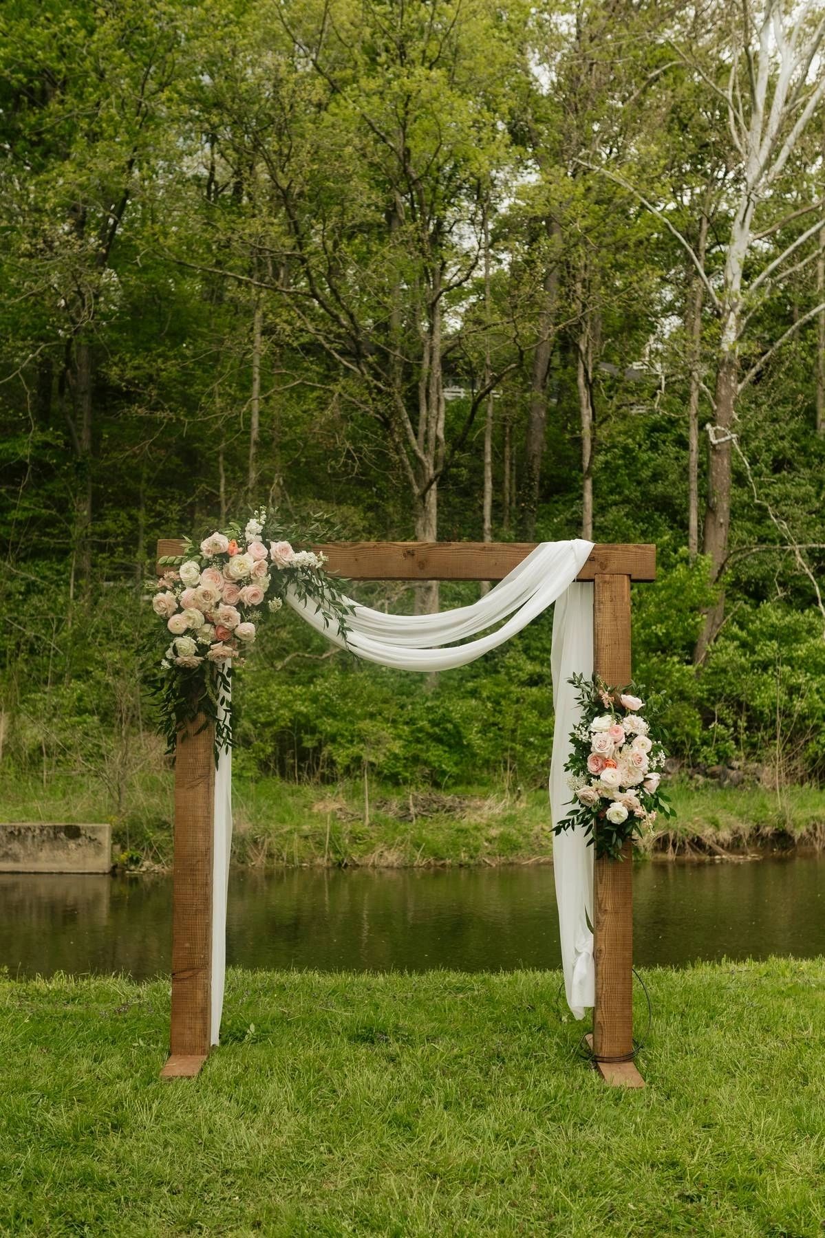Wooden ceremony arch draped in white fabric and flowers beside the pond